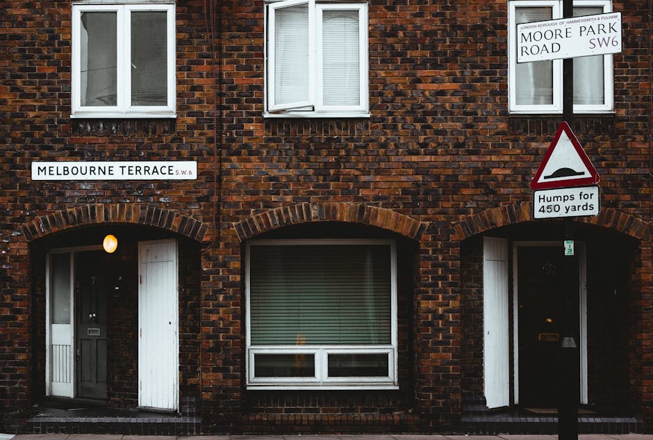 A close-up view of a brick residential building in Fulham SW6, featuring two white-framed double-hung windows with closed blinds on the upper floor. Below, on the ground floor, there are two entrances