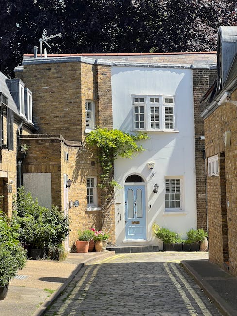 A small, charming residential garden situated in front of a white-painted building with large multi-pane windows and a blue front door. The garden features a paved pathway made of cobblestones that le