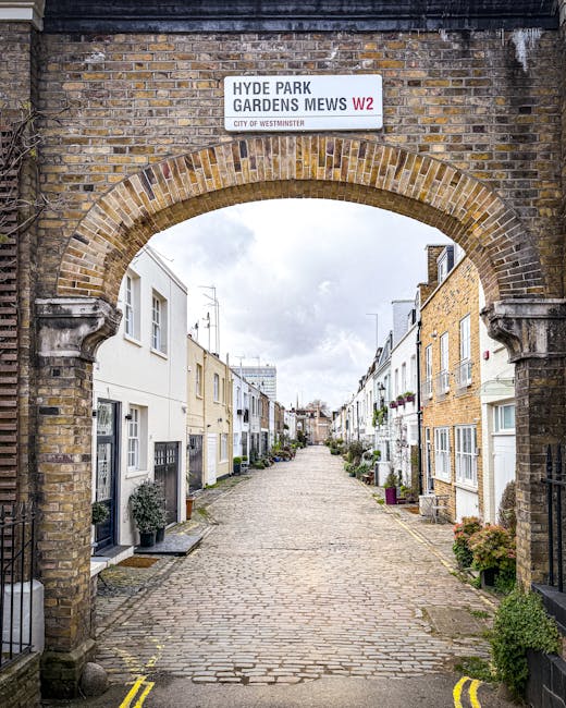 A small, charming residential garden situated in front of a white-painted building with large multi-pane windows and a blue front door. The garden features a paved pathway made of cobblestones that le